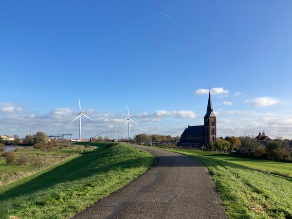 Fietspad op de dijk bij Weurt, links 2 windmolens in het zonlicht, rechts de kerktoren. In het midden verder weg de brug